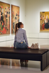 Thoughtful young Caucasian woman wearing glasses and looking at exhibition. Concept of Museum Day.