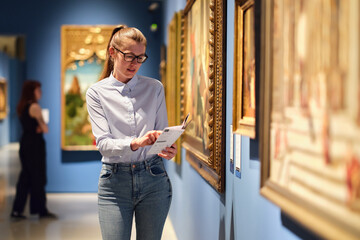Thoughtful young Caucasian woman wearing glasses and looking at exhibition. Concept of Museum Day.