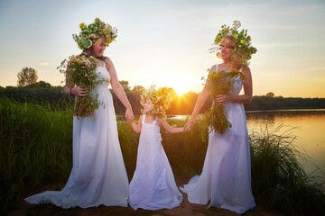 Ivan Kupala Celebration. Three Girls With Floral Wreaths by the River at Sunset. Family clad in...