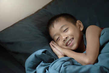 Cute boy sleeping and smiling with happy sweet dreams covered with blanket on blue bed in bedroom and morning sunlight background.
