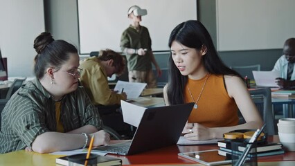 Young businesswoman with Down syndrome typing on laptop and talking with Asian female colleague while working together in office, man using VR headset in background - Powered by Adobe