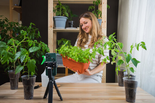 Content creator woman smiles while recording a video of herself holding a large pot of basil, surrounded by her thriving indoor garden of various potted plants.
