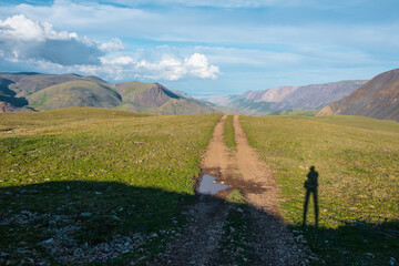 Long shadow of man on sunlit grassy green pass with dirt road with view to wide alpine valley...