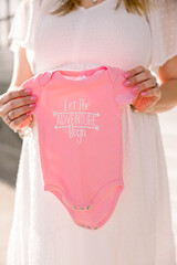 A pregnant woman wearing a white dress holds up a pink onesie that says "let the adventure begin"