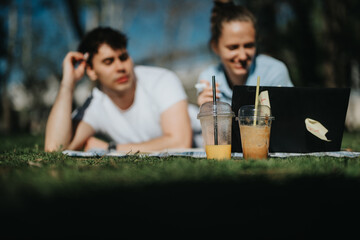 Affectionate student couple enjoying a study session in the park with cold beverages and a notebook.