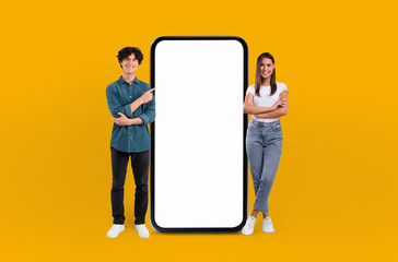 A man and a woman are standing side by side next to a large phone. The man is holding a notepad and...