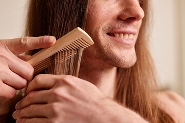Men's hair care concept. A young guy combing his long curly hair with a comb while standing in a...