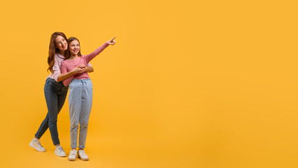 Mother and daughter dressed in casual wear, stand closely together against a vibrant yellow backdrop, pointing at copy space while they both smile brightly, sharing a lighthearted moment.