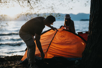 Two friends enjoy their weekend outdoors as they set up a camping tent by a beautiful lake at...
