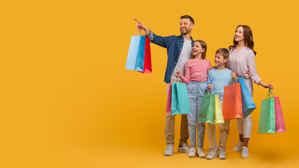 A joyful family, with two parents and two children, are standing together holding colorful shopping bags. The father points out something in the distance, copy space