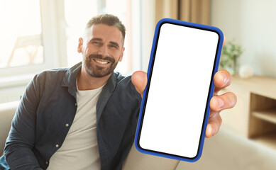 A man is seated on a couch while actively holding up a cell phone in his hand. He appears focused...