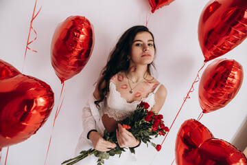 A smiling woman with kisses lipstick is holding a bouquet of red roses and standing amidst heart-shaped helium balloons, possibly celebrating a special occasion like Valentines Day
