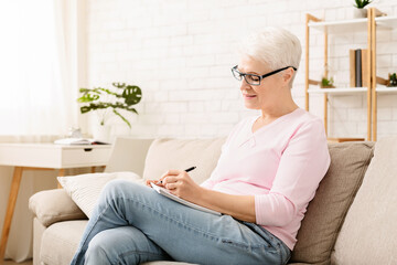 Senior woman is seated on a couch, concentrating as she writes on a piece of paper placed on her...