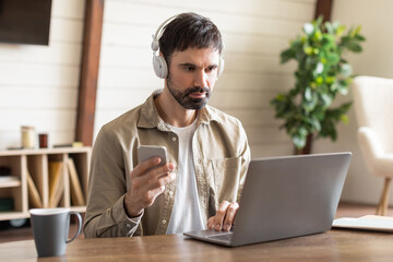 A man is seated at a wooden table, engaging attentively with his laptop while wearing headphones....