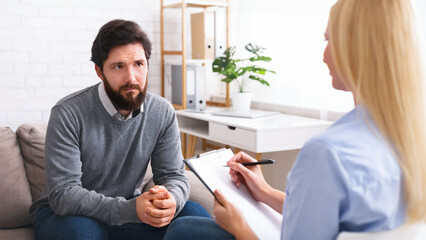 A man sits on a couch, appearing to be in earnest conversation with woman therapist who holds a notepad. They are in a well-lit, modern therapy office setting.