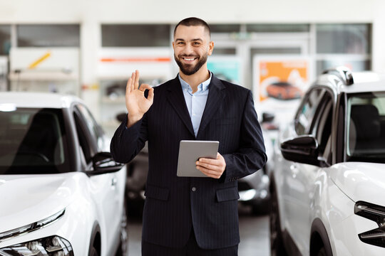 A man standing in front of a car, holding a tablet in his hand. He appears to be inspecting something on the device, with the car parked behind him.