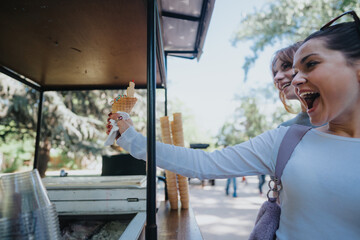 Two joyful women receiving a cone of colorful ice cream at a park stall, laughing and enjoying the...
