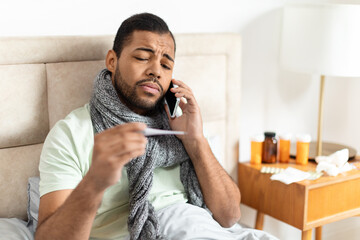 A young African American man is sitting up in bed with a gray scarf wrapped around his neck,...