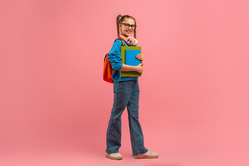 A young girl standing with a backpack hanging from her shoulder, holding a book in her hands. She...