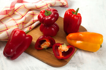 Cutting board with several whole and halves of red and yellow bell pepper and red kitchen towel on white wooden background. .