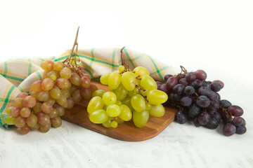 Cutting board with bunch of organic green, pink and blue grapes on white wooden background..
