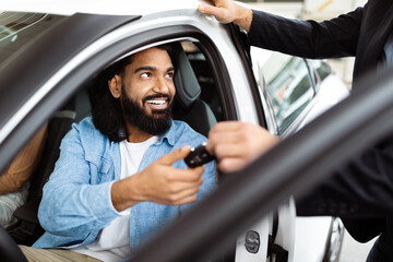 A cheerful Indian man is seated inside a car, receiving a set of car keys from a salesperson, suggesting he has possibly made a purchase or is taking a test drive