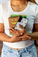  A woman is holding a cell phone and a notebook with a smiley face on it. She is wearing a white...
