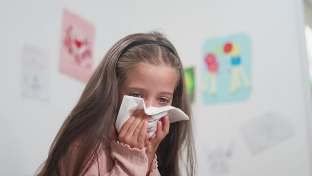 Sick girl sneezing snot into tissue paper in bedroom at home. Female child blowing runny nose is suffering from cold and flu. 