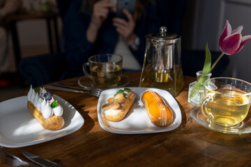 Enjoy a serene afternoon tea setting with a variety of pastries, including eclairs, served on a wooden table bathed in sunlight. The ambiance is complemented by a vibrant tulip