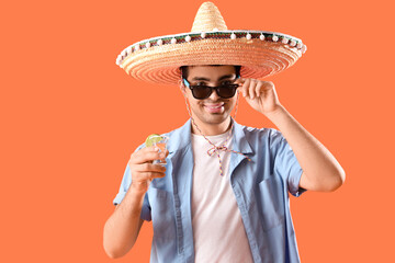 Young man in sombrero hat with tequila shot on orange background