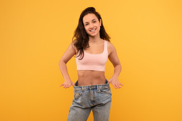 A cheerful young woman stands with hands on her hips wearing a light pink crop top and oversized...