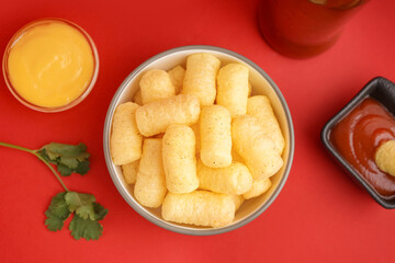 Bowl with tasty salty corn sticks and sauces on red background, closeup