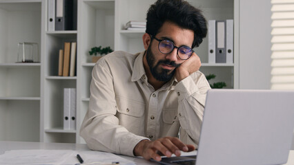Arabian Indian ethnic male tired businessman office employee boring work with laptop dreary exhausted lazy bored business man low energy overworked fatigue depression apathy burnout typing on computer