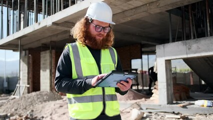 Smart engineer holding digital tablet typing with his finger on the screen of device, assessing progress of building's construction, looking around.