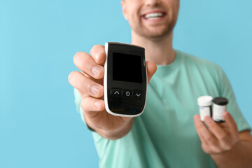 Handsome young happy diabetic man with glucometer and jars of pills on blue background, closeup