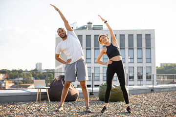 Happy sports couple making health gymnastics in morning on roof terrace of building. Positive man in white t-shirt and woman in black top doing sports stretching their arms up.