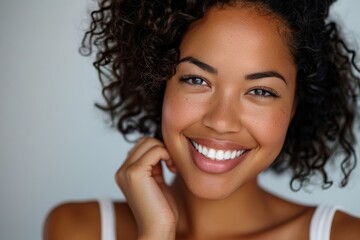 A young woman with curly hair smiling joyfully and looking into the camera, her hand delicately touching her hair
