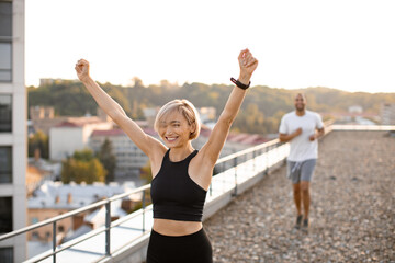 Slim happy woman in black top wins running competition from sporty man in white t-shirt during morning warm-up on roof of modern building on urban background. Healthy lifestyle concept.