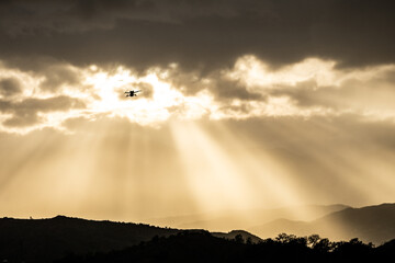 An drone in flight above mountain range with sunlight beams breaking through clouds in sky. light unmanned aircraft hovers above ground looking down at the mountainous landscape