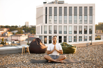 Young athletic man doing yoga at sunset on urban background. Handsome male meditating in lotus...