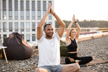 Happy healthy couple meditating on roof on urban background in morning Physically strong man and woman sitting on mats and making yoga with hands raised above their heads. . - Powered by Adobe