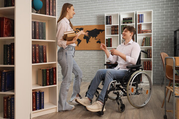 Male graduate in wheelchair talking to his friend with books at library