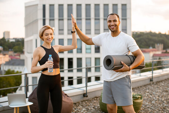 Happy sports couple giving each other high five after completing sports workout on rooftop against urban background. Cheerful husband and wife celebrate end of physical morning warm-up.