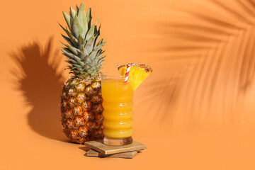 Fresh ripe pineapple with glass of tasty juice on stand against orange background