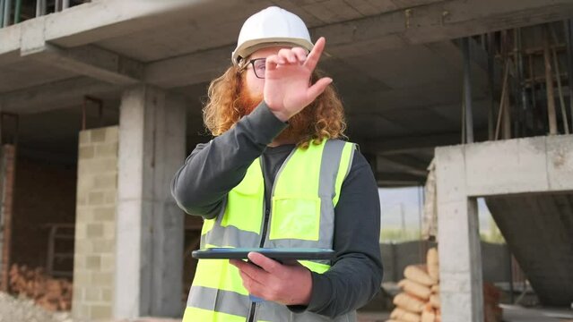 Business engineer worker man with modern hairstyle  touching an imaginary screen and pressing virtual buttons with tablet in hand, standing on construction site, outdoors.
