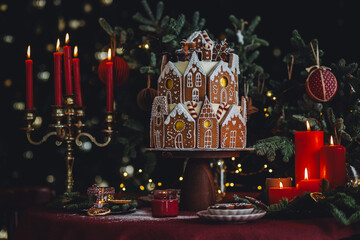 Elegant luxury Christmas table setting for family home party with homemade big gingerbread house, holiday celebration. Red tablecloth, candles, chandelier. Romantic candlelit dinner