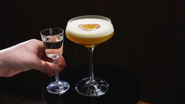 Hand of unrecognizable caucasian person placing chilled shot glass of prosecco next to porn star martini. Passion fruit flavoured cocktail with vanilla vodka and lime juice on dark background.