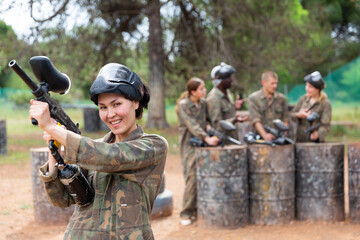Fototapeta premium Smiling asian woman paintball player in camouflage posing with gun outdoors
