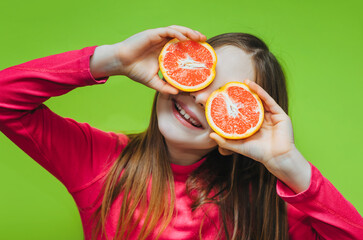 Little cute funny girl uses grapefruit for eyes and has fun in the studio on a green background. Concept of citrus diet, fruits and vitamins.