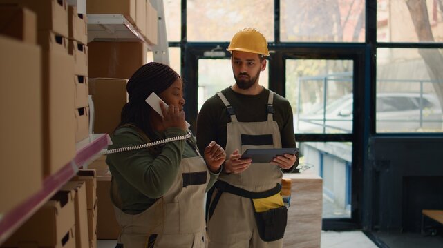 African american head of logistics and employee preparing warehouse orders for delivery, using tablet to verify shipping informations, receiving telephone call while scanning labels in depository